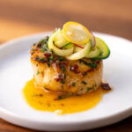 closeup of a single scallop plated with melted chipotle butter and a small garnish of zucchini and radish on top.
